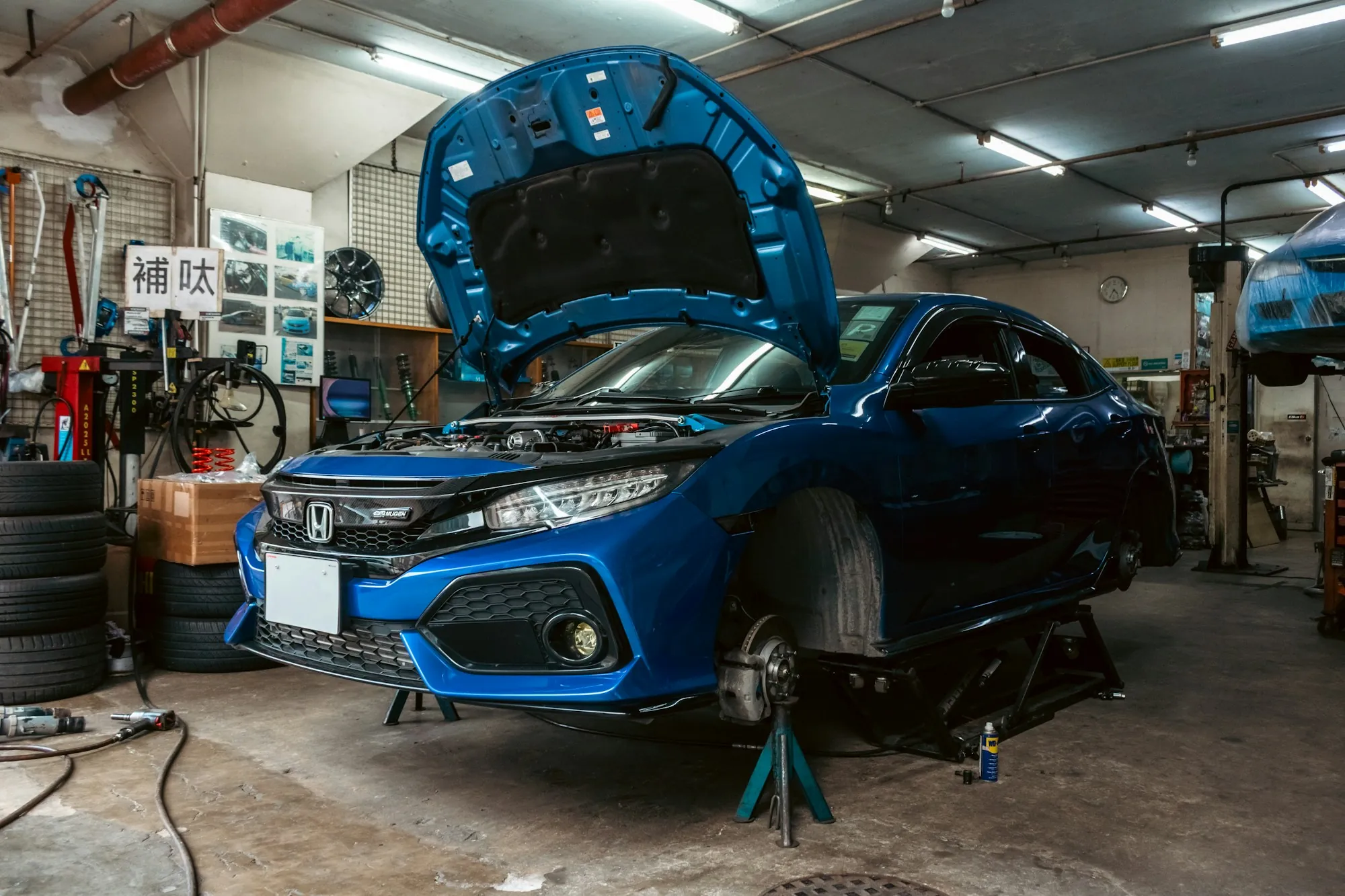 Blue hatchback with its hood up inside a busy repair shop